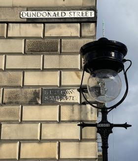 White letters on a black background, painted directly onto masonry and reading DUNCAN STREET. Above, a more recent sign reading DUNDONALD STREET. In the foreground a period-style lamppost.