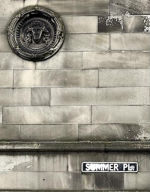 Miserable-looking soot encrusted face at centre of carved medallion on side of building.